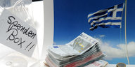Vor einer griechischen Flagge stapeln sich Geldscheine, daneben eine Kiste mit der Aufschrift "Spendenbox".