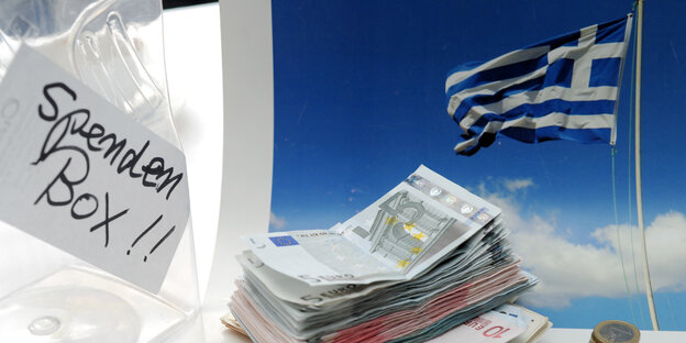 Vor einer griechischen Flagge stapeln sich Geldscheine, daneben eine Kiste mit der Aufschrift "Spendenbox". Vor einer griechischen Flagge stapeln sich Geldscheine, daneben eine Kiste mit der Aufschrift "Spendenbox".