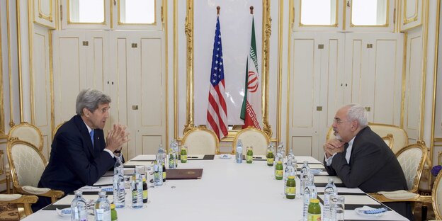 zwei Männer sitzen sich an einem gedeckten Tisch gegenüber, dahinter die Fahnen von USA und Iran zwei Männer sitzen sich an einem gedeckten Tisch gegenüber, dahinter die Fahnen von USA und Iran