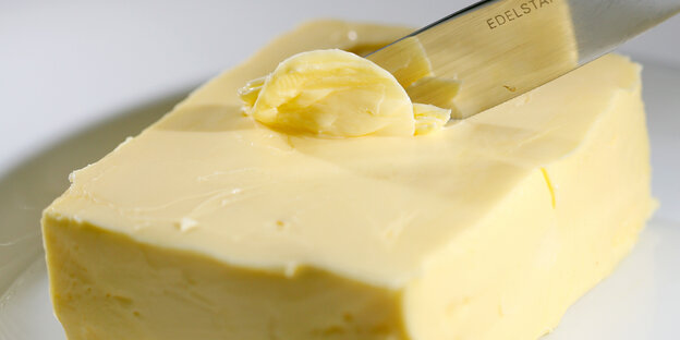 Wir sehen ein Stück Butter auf einem weißen Teller und ein Messer, das sich in die Butter gräbt. Wir sehen ein Stück Butter auf einem weißen Teller und ein Messer, das sich in die Butter gräbt.