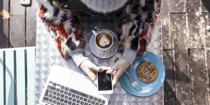 Eine Frau sitzt an einem Tisch und hält ein Smartphone in der Hand - auf dem Tisch ein Kaffee mit Herz im Schaum, ein Cookie und ein Laptop