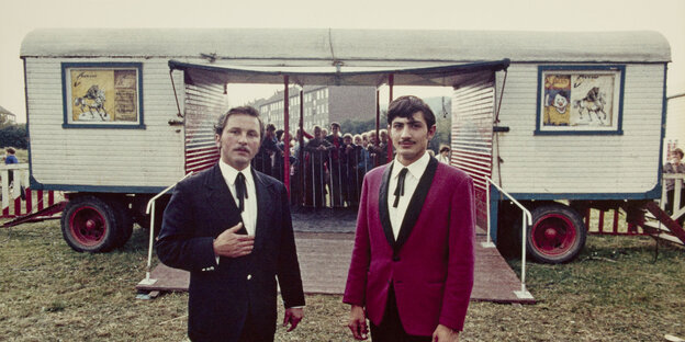 Zwei Männer in Anzügen stehen vor einer Zirkuskasse. Im Hintergrund stehen Menschen an, dahinter ist in der Ferne ein Hochhaus zu sehen