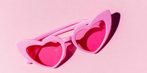 Auf einem pinken Untergrund liegt eine pinke Brille, deren Gläser herzförmig und rot gefärbt sind