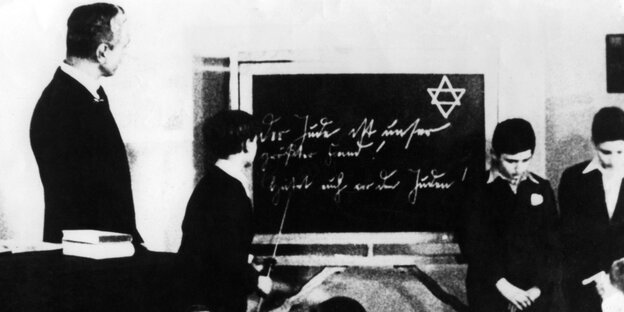 Eine Schulklasse zur NS-Zeit. Ein Schüler steht vor einer Tafel, auf der ein Davidstern zu sehen ist. Eine Schulklasse zur NS-Zeit. Ein Schüler steht vor einer Tafel, auf der ein Davidstern zu sehen ist.