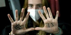 Junge Frau mit Atemmaske hat auf die Handfläche der rechten Hand ein Auge gemalt. Auf der Linken steht 1,5