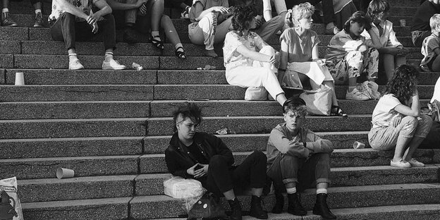 Schwarz-weiß-Fotografie. Zwei Punker sitzen auf einer Treppe. Umringt von Menschen. Schwarz-weiß-Fotografie. Zwei Punker sitzen auf einer Treppe. Umringt von Menschen.