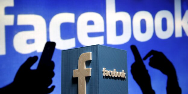 Man sieht den Facebook-Schriftzug groß im Hintergrund. Im Vordergrund Schatten von Händen, die auf Smartphones tippen. Man sieht den Facebook-Schriftzug groß im Hintergrund. Im Vordergrund Schatten von Händen, die auf Smartphones tippen.