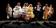 Sieben junge Schauspieler:innen laufen lachend über eine Theaterbühne auf die Kamera zu, sie tragen Kleidung aus dem 19. Jahrundert, auf ihren Körpern ist Schrift projizieret. Es ist zu lesen "Utopie für Anfänger"