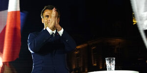 Der französische Präsident Emmanuel Macron steht flankiert von einer französischen Fahne mit in einer Dankbarkeitsgeste gefalteten Händen im Scheinwerferlicht.