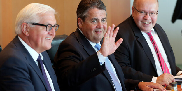 Drei Männer nebeneinander am Tisch, einer hebt die Hand Drei Männer nebeneinander am Tisch, einer hebt die Hand