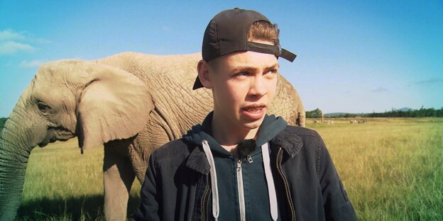 Ein Jugendlicher mit umgedrehtem Baseballcap und Zahnspange schaut leicht zur Seite. Im Hintergrund ein Elefant