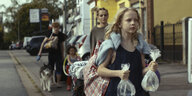 Szene aus „Petite nature“: Eine Familie läuft mit viel Gepäck und einem Hund eine Straße entlang, vorne rechts im Bild ist der zehnjährige Johnny zu sehen. Er trägt zwei mit Wasser gefüllte Plastikbeutel in der Hand, in denen Fische schwimmen, und eine g