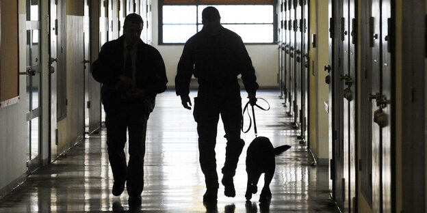 Zwei Männer in Uniform gehen mit einem Hund an der Leine über einen Zellenflur