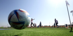 Der WM Ball fliegt über en Platz, im Hintergrund Fussballer die trainieren