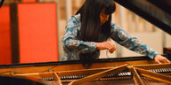 Satoko Fujii steht an einem an der Oberseite geöffneten Steinway Flügel und greifzt mit einer Hand ins Innere. Die Pianistin trägt ein türkis gemustertes Hemd und lange Haare