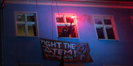 Ein Vermummter hält aus einem Fenster ein begalo. Unter ihm auf einem Transpi steht "Fight the system"