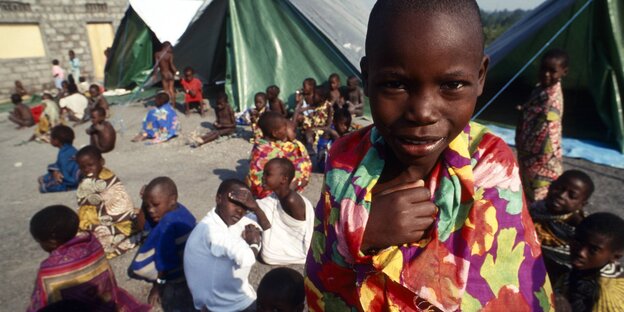 Ruandische Flüchtlingskinder. Eines steht im Vordergrund und schaut in die Kamera. Weitere, vor Zelten sitzende Kinder im Hintergrund. Ruandische Flüchtlingskinder. Eines steht im Vordergrund und schaut in die Kamera. Weitere, vor Zelten sitzende Kinder im Hintergrund.