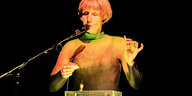 Die Klangkünstlerin Ute Wassermann steht auf einer Bühne und singt in ein Mikrofon. Sie hält eine Vogelpfeife in der Hand. Vor ihr steht ein Tisch, auf dem sich ein quadratischer, transparenter Behälter mit Wasser befindet. Wassermann hält ein Kontaktmikr