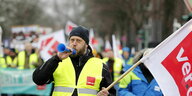 Ein Mann in einer Weste der Gewerkschaft Verdi und einer Gewerkschaftsfahne steht vor zahlreichenden Streikenden und pustet in eine Tröte