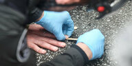 Polizist mit blauen Gummihandschuhen löst mit einem Holzspachtel die festgeklebten Hände eines Aktivisten von der Straße.