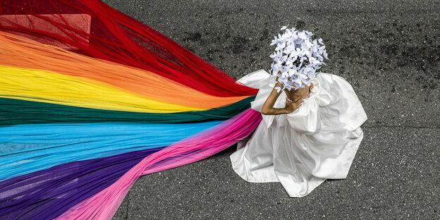 A person with a white skirt and white flowers on his head wears a rainbow veil