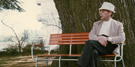 Simenon sitzt mit hellem Hut auf einer roten Parkbank vor einem dicken Baumstamm