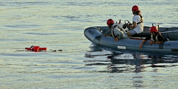 A person in an orange life jacket in the water and 3 people with helmets in a dinghy with a motor from the organization Mare Liberum