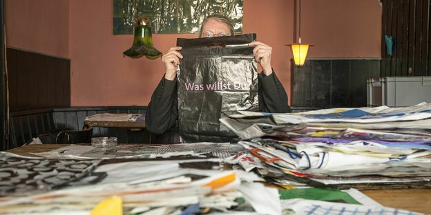 Jürgen Francke sitzt in einer Kneipe vor vielen Plastiktüten, sein Gesicht halb verdeckt von einer mit der Aufschrift: „Was willst du?“