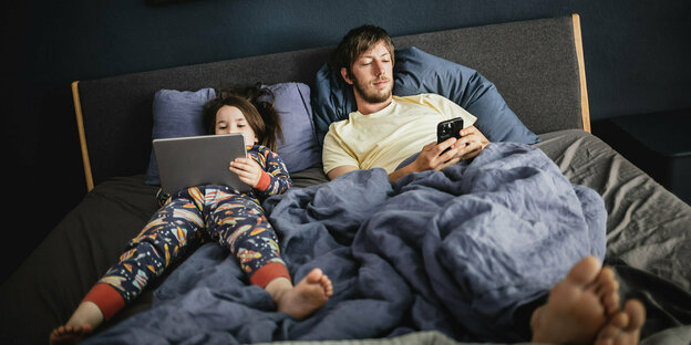 A man is lying in bed with a smartphone, next to him is a child with a tablet and colorful pajamas
