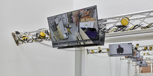 Exhibition view: Screens showing surveillance camera footage are attached to crossbeams