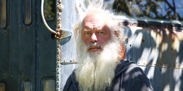An old man with a white beard and tousled hair looks into the camera