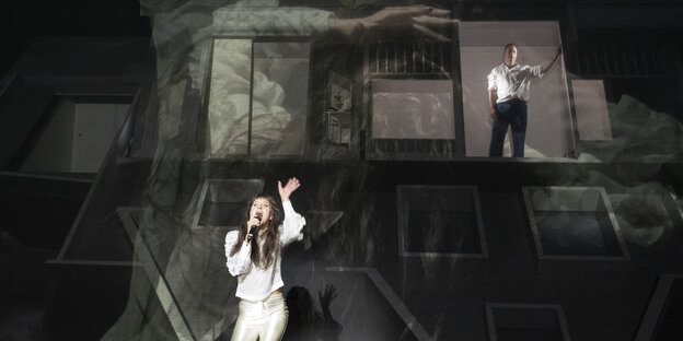 Eine dunkelhaarige Frau singt inbrünstig inmitten eines pompösen Bühnenbilds . Rechts hinter ihr, erhöht, steht ein Mann im Fensterrahmen.