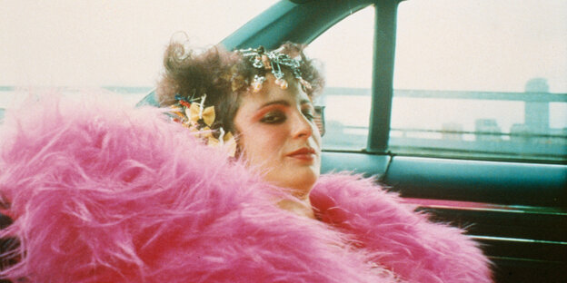 Eine Frau mit lockigen Haaren, auffällig geschminkt und in rosarote Federn gehüllt