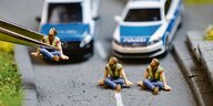 Knetfigürchen Letzte Generation auf einer Straße, dahinter zwei Polizeiwagen