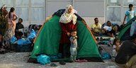 Eine Frau mit einem kleinen Kind steht vor einem Zelt, dahinter viele Menschen, die auf dem Boden kauern