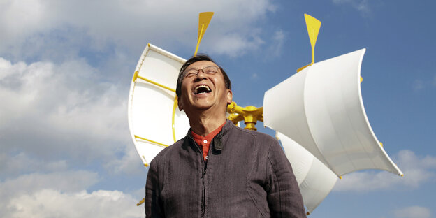 Der Künstler Susumu Shingu steht vor einer kinetischen Skulptur aus gelben Stahlträgern und weißen Stoffsegeln. Dahinter ist der blaue Himmel mit Wolken zu sehen. Der Künstler trägt eine graue Jacke, darunter ein orangenes Hemd und eine Brille. Er hebt den Kopf in Richtung Hiummel und lacht fröhlich mit offenem Mund.