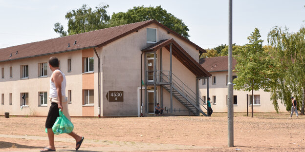 Mann schlappt mit Plastiktüte über kargen Hof an einem Haus vorbei