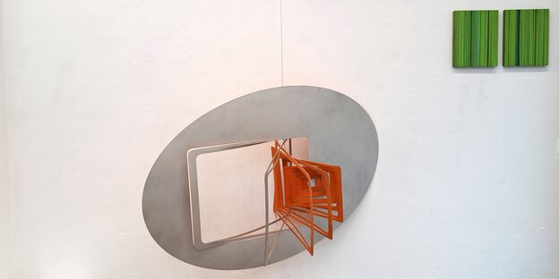 Blick in die Ausstellung: An der Wand hängt links eine ovalförmige Aluminiumskulptur von Susanne Specht, aus der mehrere rechteckige und orange gestrichende Rahmen herausragen. Recht über der Skulptur hängen zwei grüne Bilder von beate maria wörz. Papierb
