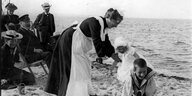 Eine Kinderfrau in Schürze spielt mit zwei Kindern am Strand, im Hintergrund schauen zwei Männer und zwei Frauen zu