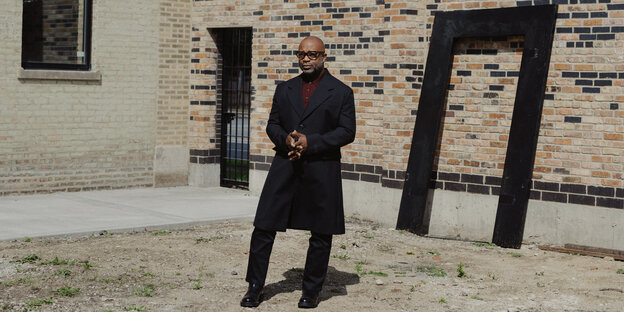 Theaster Gates is standing in front of a building, wearing a long black coat and a burgundy shirt