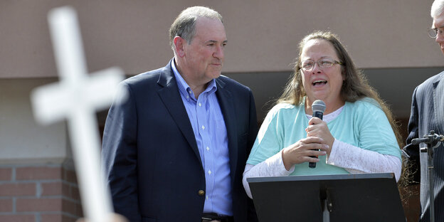 Kim Davis spricht in ein Mikrofon, neben ihr der republikanische Präsidenschaftskandidat Huckabee. In der Menge halten Menschen Kreuze hoch.