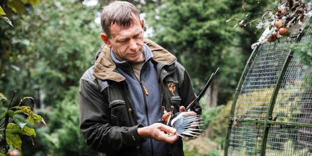 Ein Ornitologe steht neben einer Voliere und hält einen Vogel in den Händen