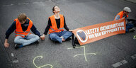 Drei Menschen von der Letzten Generation sitzen in Warnwesten und mit einem Transparent auf einer Straße.
