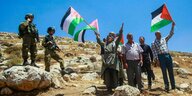 Männer schwenken pälästinensische Flaggen auf einem kargen Hügel, beobachtet von zwei schwer bewaffneten israelischen Soldaten
