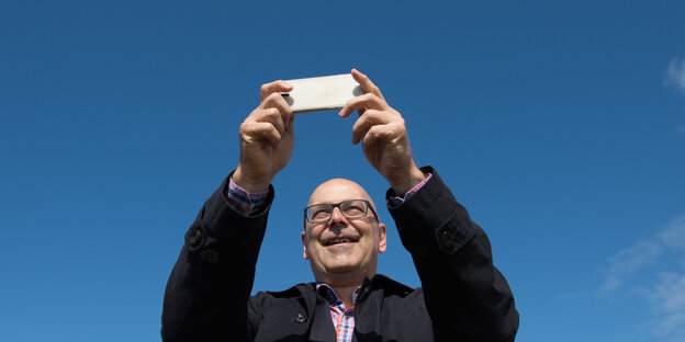 Thorsten Albig hält vor blauem Himmel ein Smartphone mit beiden Händen hoch Thorsten Albig hält vor blauem Himmel ein Smartphone mit beiden Händen hoch
