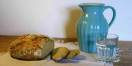 Auf einem Tisch mit beiger Decke liegt ein aufgeschnittener Laib Brot, eine blauer Krug und ein Glas mit Wasser