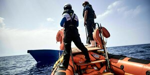 Zwei Männer stehen auf einem Schlauchboot mit dem sie sich einem leeren Boot auf hoher See nähern. Auf deren Tauchanzug steht SOS Mediterranee