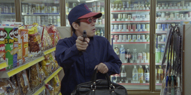 Ein junger Mann steht mit großer Tasche und einer Waffe zwischen Regalen im Supermarkt