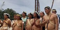 Menschen mit nacktem Oberkörper und Blumenschmuck im Haar stehen vor einem Erdölturm
