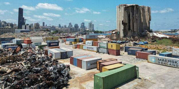 Von links nach rechts sind auf dem Bild zu sehen: Ein Haufen Altmetallschrott, eine Reihe bunter Schiffscontainer, ein zerstörtes Gebäude